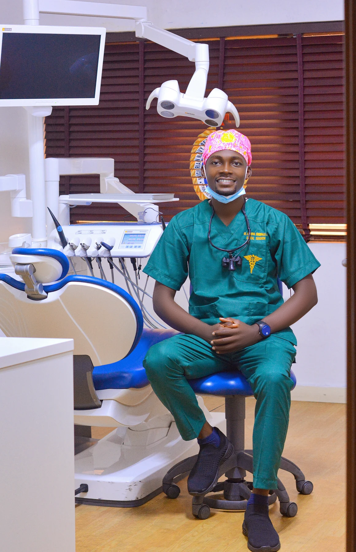 Dr. Iortyom, Aondongu Theophilus in surgical scrubs and dental loupes beside a treatment chair in a dental surgery
